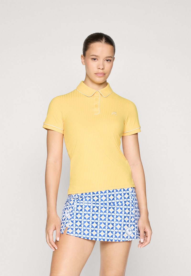 Polo shirt giallo a coste con colletto a punta, accentuato da bordi bianchi, abbinato a una gonna blu a fantasia con design circolari.