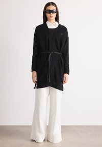 Μαύρο πλεκτό πουλόβερ με ζώνη στη μέση, featuring ριμπ μανσέτες. Συνδυασμένο με λευκά παντελόνια σε φαρδύ στιλ και κομψά μαύρα γυαλιά ηλίου.