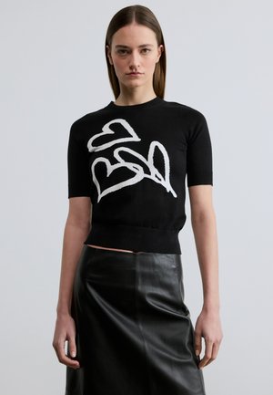 Žena v černém tričku s krátkými rukávy a bílými vzory srdcí a v černé kožené sukni stojí před jednoduchým světlým pozadím.