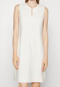 Robe sans manches, jusqu'aux genoux, de couleur crème clair, avec un petit col en V et un tissu lisse et structuré avec des coutures latérales.