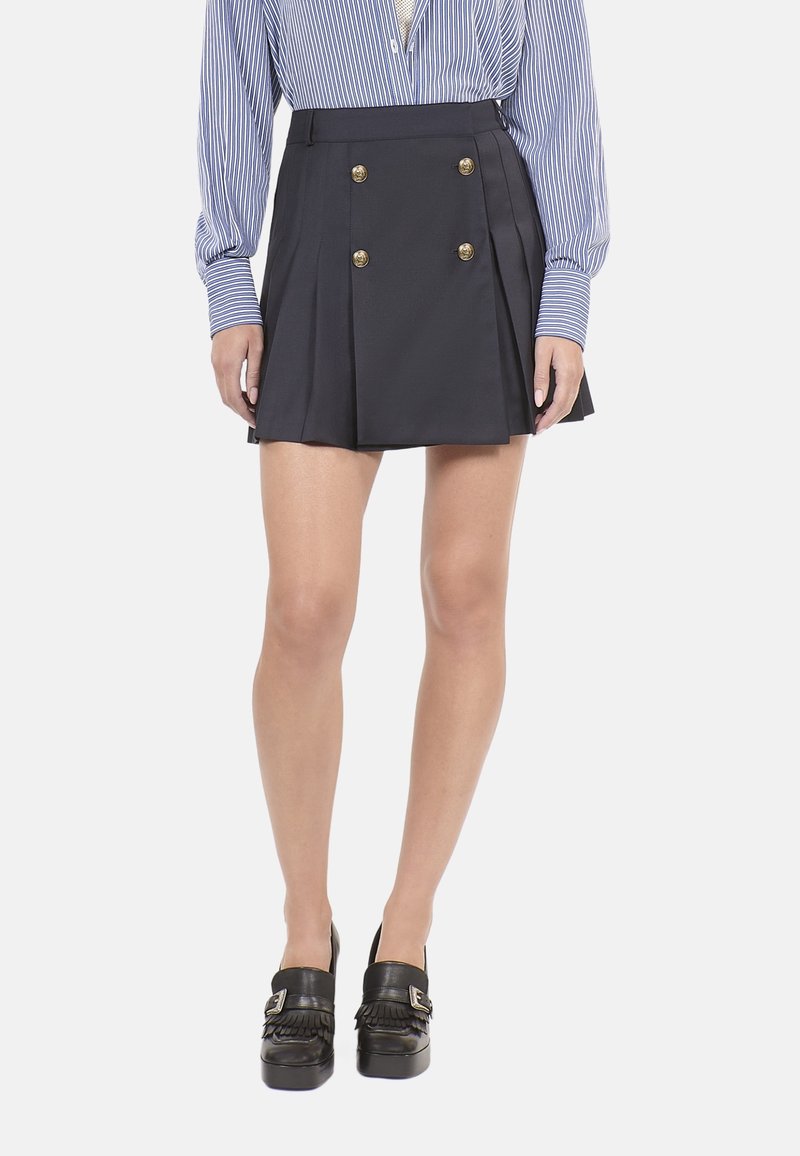 Marineblaue Faltenrock mit doppelten goldenen Knöpfen, aus einem glatten Stoff gefertigt. Kombiniert mit einer gestreiften Bluse und schwarzen Plateauschuhen.