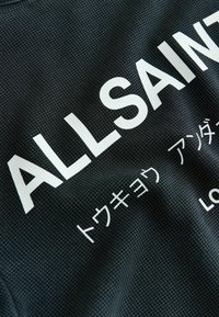 Sort tekstureret stof med stor hvid tekst "ALL SAIN" og mindre japanske tegn nedenunder, delvist synlige og let krøllede.