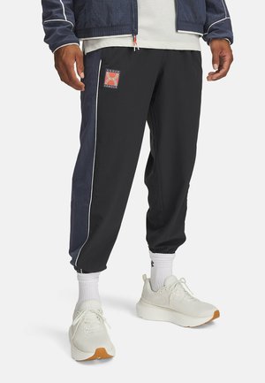 Man draagt zwarte en marineblauwe trainingsbroek met witte biezen, witte sokken en witte sportschoenen, staand tegen een effen achtergrond.