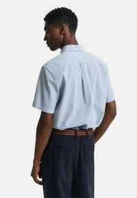 Kurzärmliges Hemd mit Knopfleiste in einem blau-weißen Streifenmuster, ausgestattet mit einer Rückeneinlage und Falten. Kombiniert mit dunklen Hosen und einem geflochtenen Gürtel.