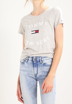 Kobieta ubrana w jasnoszarą koszulkę Tommy New York i jasnoniebieskie dżinsy z wysokim stanem, stojąca z jedną ręką swobodnie opuszczoną wzdłuż ciała.