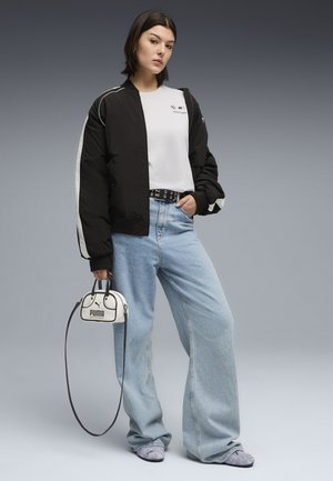 Jeune femme portant une veste noire et blanche, un t-shirt blanc, un jean large bleu clair, des chaussures grises, tenant un petit sac à main blanc Puma avec une longue bandoulière.