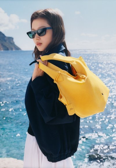 Νεαρή γυναίκα με γυαλιά ηλίου στέκεται δίπλα στη θάλασσα, φοράει μπλε σκούρο μπουφάν και λευκή φούστα, κρατώντας μια μεγάλη, φωτεινή κίτρινη δερμάτινη τσάντα στον ώμο.