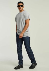 Szara koszulka z krótkim rękawem z podwiniętymi mankietami, niebieskie dżinsy oraz czarne buty. Ubranie charakteryzuje się minimalnymi akcentami marki.