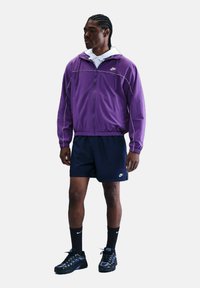 Giacca in nylon viola con accenti bianchi, indossata sopra un hoodie bianco. Pantaloni corti blu navy, calze nere e sneaker nere completano l'outfit atletico.