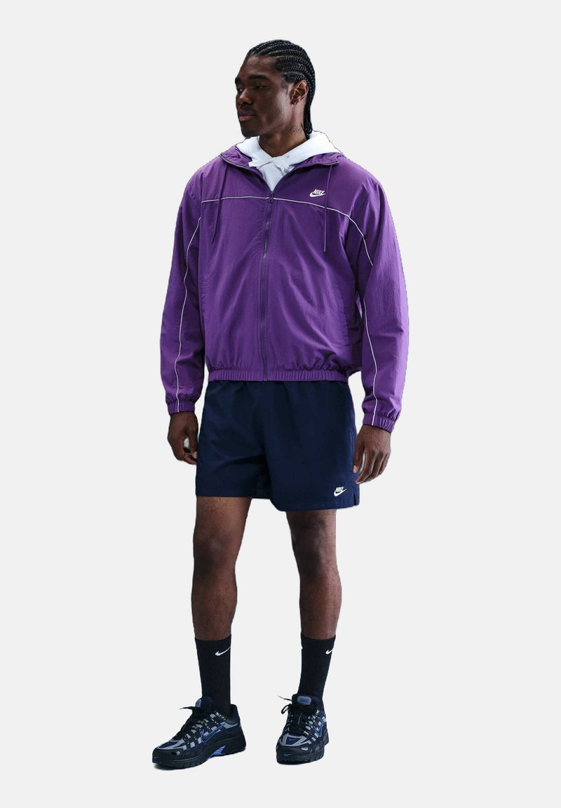 Giacca in nylon viola con accenti bianchi, indossata sopra un hoodie bianco. Pantaloni corti blu navy, calze nere e sneaker nere completano l'outfit atletico.