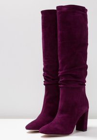 Botas de gamuza moradas hasta la rodilla con un diseño holgado, punta afilada y un tacón alto grueso. Textura suave sin herrajes visibles.