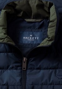 Veste matelassée bleu marine avec col intérieur vert olive, fermeture éclair centrale et étiquette Hackett London à l'intérieur du col.