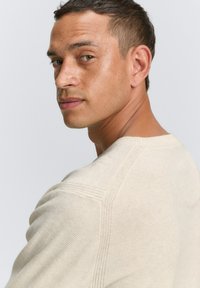 Hellbeige strukturierter Pullover mit gerippten Akzenten, rundem Ausschnitt und figurnaher Passform, an einer Person seitlich dargestellt.