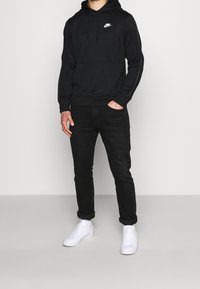 Schwarzer Hoodie mit Fronttasche und weißem Nike-Logo, kombiniert mit schwarzen Jeans und weißen Sneakers. Mit Kordeln und strukturierten Ärmeln ausgestattet.