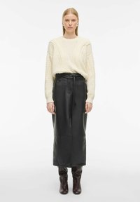 Pull en maille torsadée crème associé à une jupe midi en cuir noir, avec une taille ceinturée, et des bottes hautes marron foncé. Texture lisse.