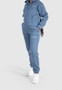 Sudadera corta azul con bolsillo frontal y cremallera, combinada con pantalones joggers a juego con puños elásticos. Acentuada con branding en blanco.