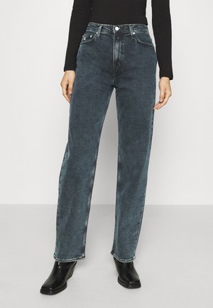 Osoba ubrana w czarne, wyblakłe jeansy z wysokim stanem i prostymi nogawkami oraz czarne botki za kostkę, a także czarną bluzkę z długim rękawem, stojąca na jednolitym tle.