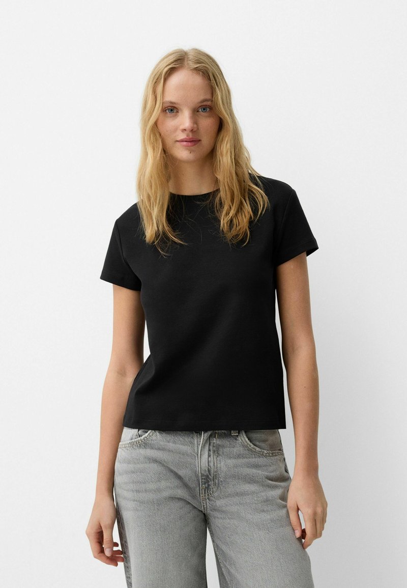 Tricou negru cu maneci scurte, din bumbac, cu un croi drept și guler rotund. Asortat cu blugi de denim gri deschis.