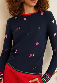 Modella con capelli ricci che indossa un maglione blu navy decorato con cuori, stelle e note musicali ricamate in colori vivaci, abbinato a pantaloni rossi.