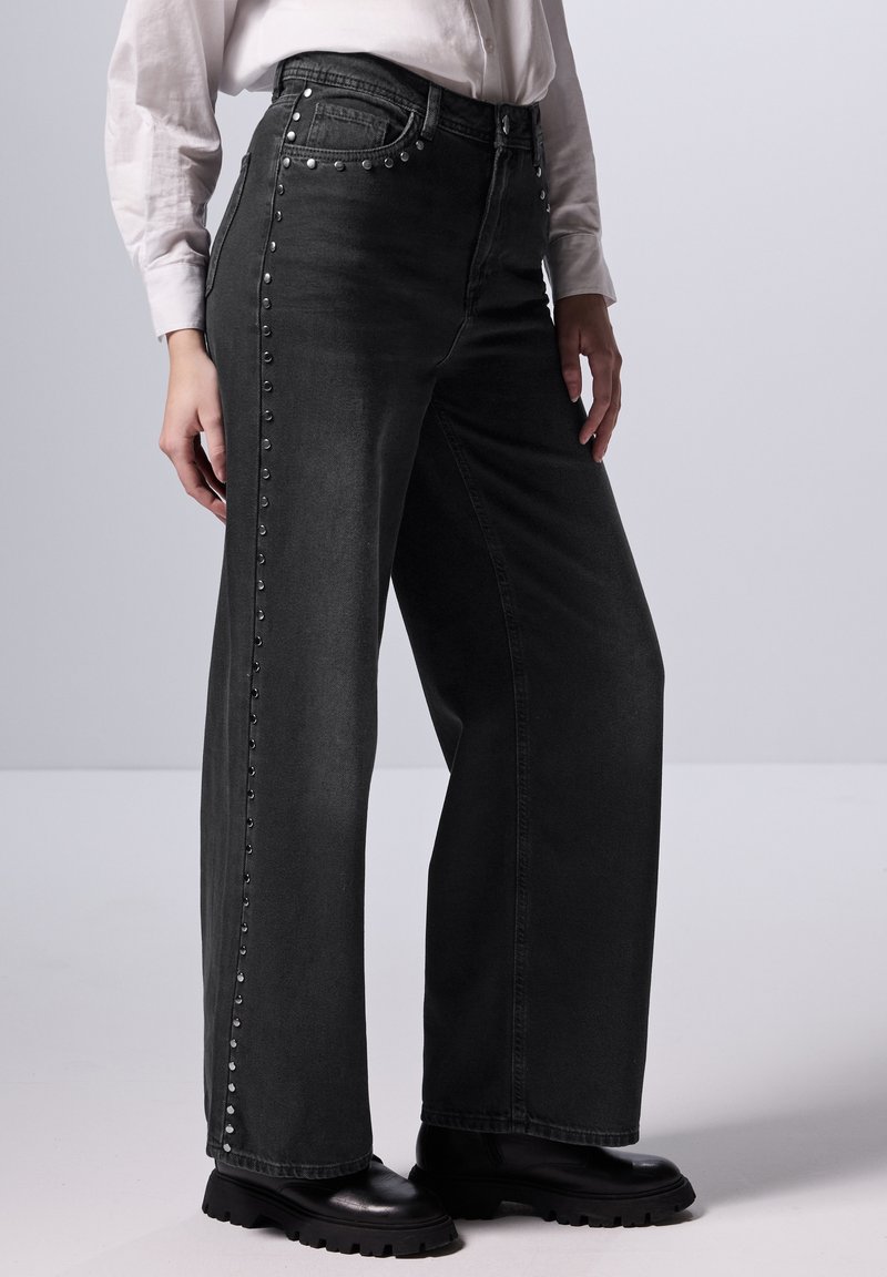 Vaqueros negros de tiro alto y pierna ancha con remaches plateados a lo largo de las costuras externas. Fabricados en denim con una textura suave. Se combinan bien con botas negras.