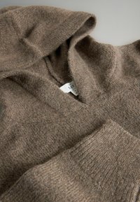 Miękki brązowy sweter z dzianiny wełnianej z kapturem i ściągaczami na rękawach, ułożony płasko na jasnej powierzchni.