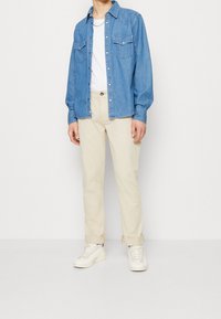 Light blue denim shirt with two chest pockets, white T-shirt underneath, cream-colored pants, and white sneakers. Minimalistic design.