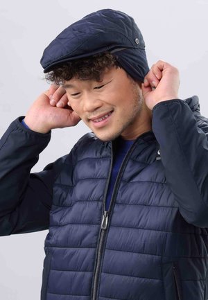Casquette matelassée bleu marine à visière plate, dotée de fixations sur les côtés, portée par une personne dans une doudoune bleu marine assortie avec fermeture zippée.