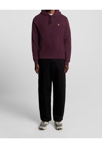 Sudadera burdeos con un bolsillo frontal y un logo amarillo, combinada con pantalones negros de corte holgado y zapatillas grises con estampado.