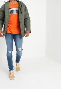 Grön parkasjacka, orange t-shirt med tryck, slitna ljusblå jeans och gula sneakers, som visar en avslappnad stadstilsstil med tydliga färger.
