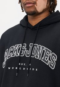 Sort hættejakke med en frontlomme, der har hvid grafisk tekst "JACK&JONES EST. 1990 WORLDWIDE." Fremstillet af bomuldsstof og med justerbar snor.