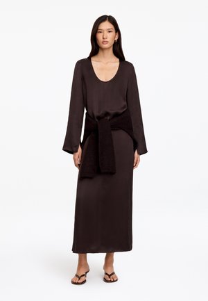 Femme portant une longue robe noire à encolure ronde, un pull noir noué à la taille et des sandales noires, debout sur un fond blanc.