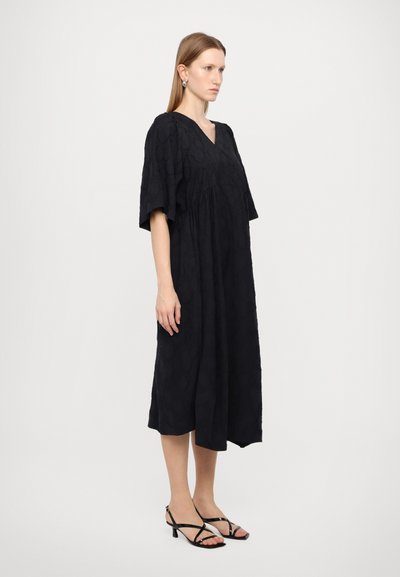 Zwarte jurk tot de knie met een V-hals, korte mouwen, een textuurstof en een geplooide taille. Gemaakt met zwarte sandaaltjes met bandjes.