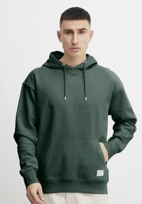 Sudadera con capucha verde, hecha de tela suave, que cuenta con un amplio bolsillo frontal, cordones y una etiqueta sutil en el dobladillo.