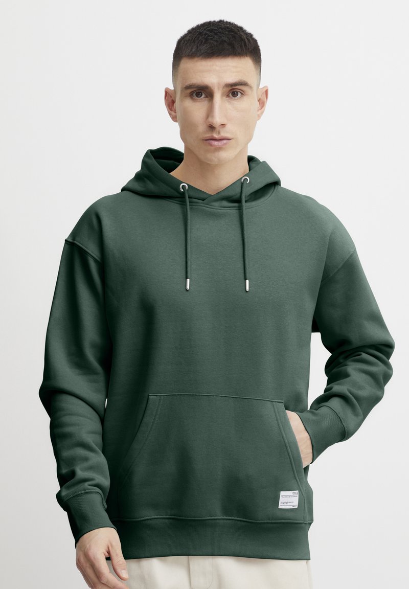 Sudadera con capucha verde, hecha de tela suave, que cuenta con un amplio bolsillo frontal, cordones y una etiqueta sutil en el dobladillo.