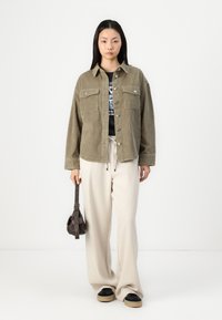 Olive corduroy jacket with two chest pockets, button front, and cuffed sleeves, paired with a black graphic tee and cream wide-leg pants.