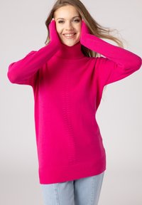 Maglione a collo alto fucsia con polsini a coste, dotato di una cucitura centrale e una vestibilità morbida. Indossato con jeans azzurri chiari.