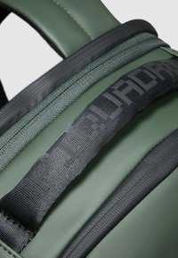 Mochila verde con material texturizado, que cuenta con una correa negra con logo en relieve y un detalle de cremallera lisa. La tela incluye acentos tejidos.