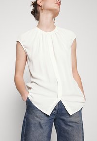 Femme portant un chemisier blanc sans manches avec un col plissé et une fente à l'avant, associé à un jean bleu, mains dans les poches.