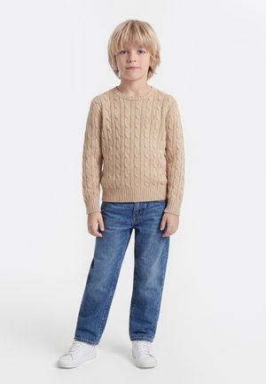 Enfant blond debout, portant un pull beige en maille torsadée, un jean bleu et des baskets blanches, face à un fond uni.