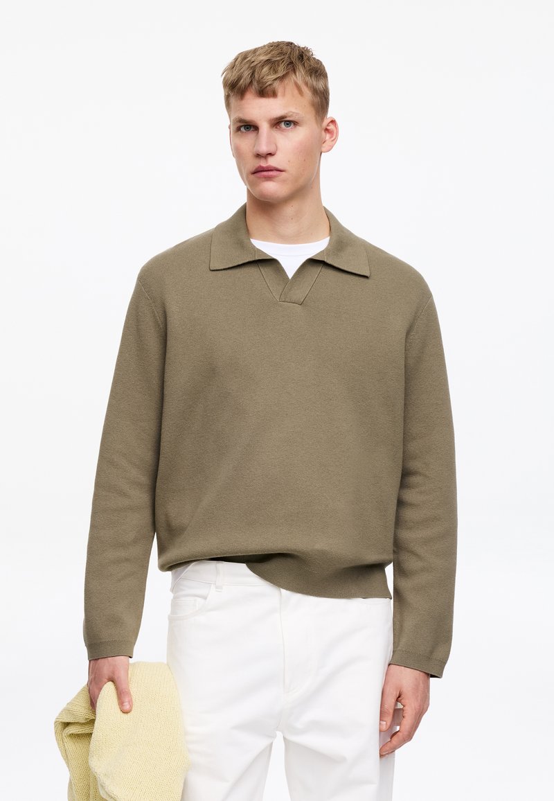 Jeune homme portant un pull col olive et un pantalon blanc, tenant un vêtement jaune en maille plié dans sa main gauche.