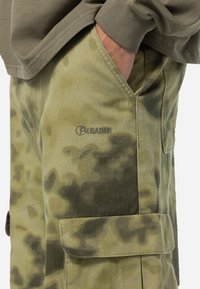 Pantalones cortos cargo de camuflaje verde hechos de algodón. Presenta bolsillos y un logo bordado de "Pegador". Tejido texturizado con patrones de manchas en varias tonalidades.