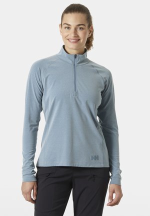 Kobieta w jasnoniebieskim topie z długim rękawem zapinanym na zamek i czarnych spodniach, uśmiechająca się, stojąca na tle jednolitego białego tła.