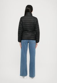 Zwarte gewatteerde pufferjas met een hoge kraag en een gestructureerde afwerking, gecombineerd met lichtblauwe wijde jeans en witte sneakers.
