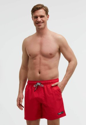 Lächelnder, oberkörperfreier Mann mit kurzen braunen Haaren, der rote Badeshorts mit einer schwarz-weißen Kordel trägt, vor einem einfarbigen hellen Hintergrund.