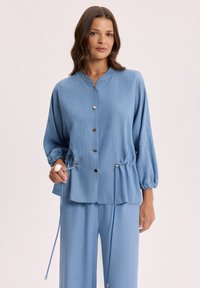 Kobieta regulująca sznurek w luźnej, jasnoniebieskiej kurtce na guziki z długimi rękawami, w zestawie z dopasowanymi szerokimi spodniami na jednolitym tle.