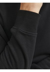 Gros plan d'une manche de sweat-shirt noire avec un poignet côtelé portée sur le bras d'une personne sur un fond sombre.