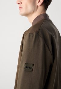 Brown bomber jacket with a smooth texture, featuring a darker fabric on the shoulders and a black square logo patch on the sleeve.