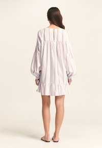 Donna in un vestito bianco con strisce verticali rosse e blu, maniche lunghe a palloncino e orlo arricciato, in piedi di spalle su uno sfondo neutro.