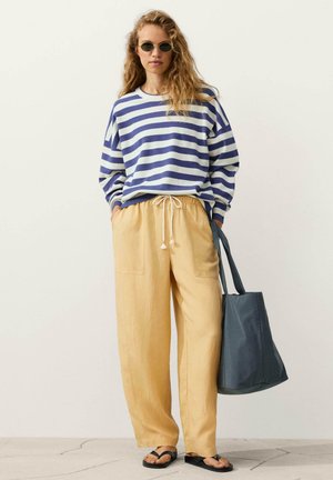 Nő, kék-fehér csíkos hosszú ujjú pólóban, sárga húzózsinóros nadrágban, fekete szandálban, napszemüveggel, egy nagy sötétszürke vászontáskát tart.