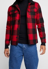 Rød og sort flanneljakke med fleecekrave, frontlukning med knapper og to brystlommer. Bæret over en sort højhalset sweater.